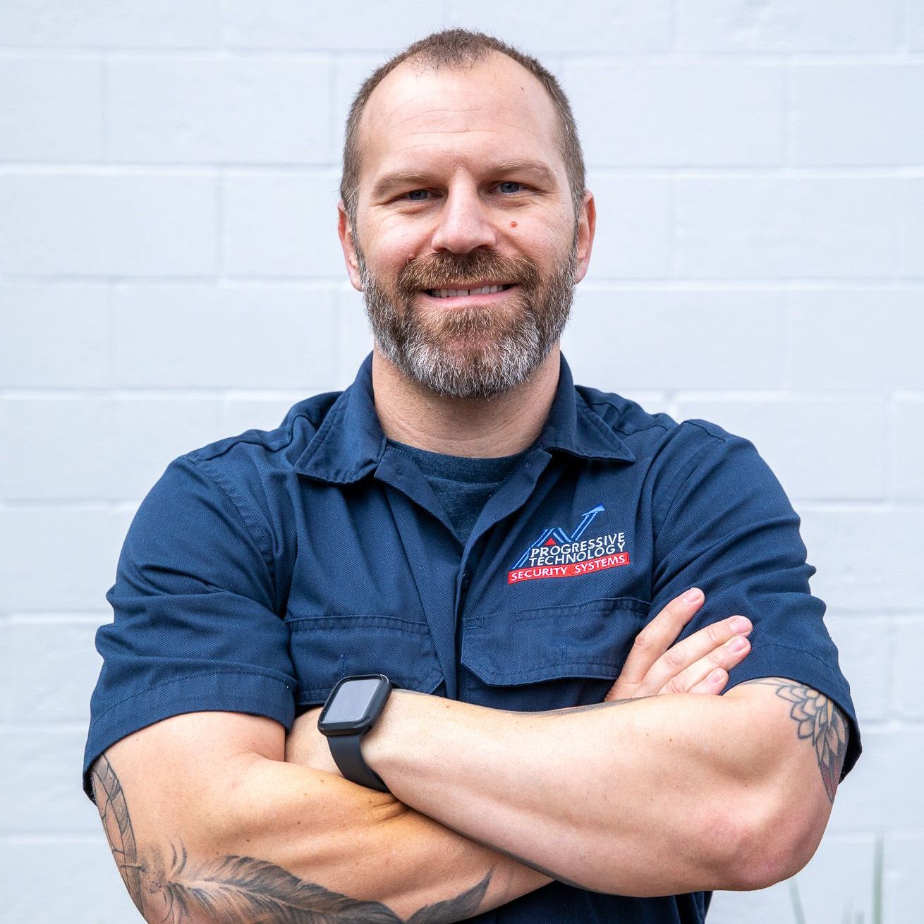 Man with crossed arms, wearing a blue work shirt, smiling in front of a white brick wall.