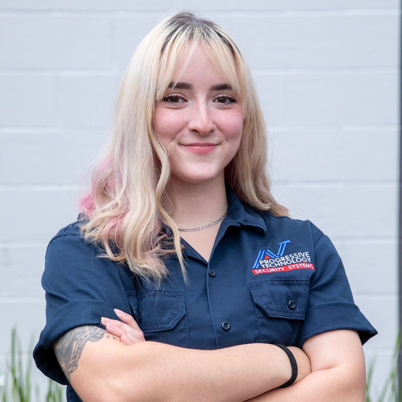 Woman with blonde hair, arms crossed, wearing a blue work shirt with a logo, smiling.