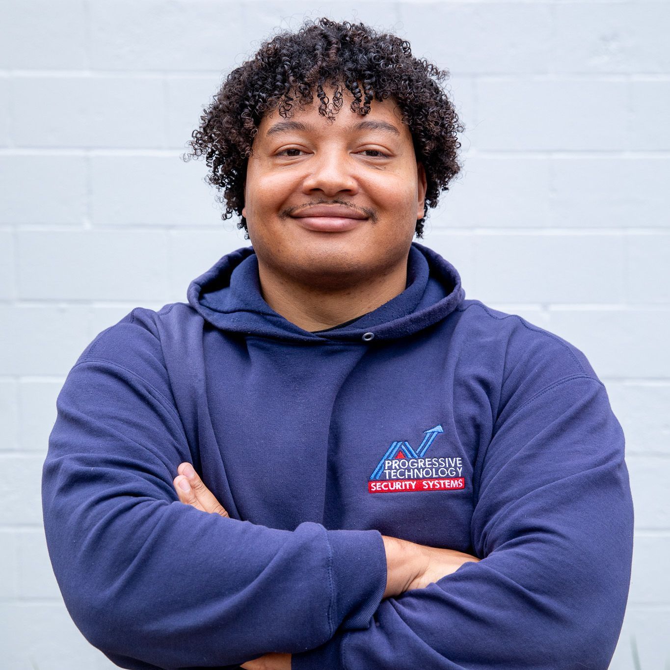 Man with curly hair in blue hoodie, arms crossed, smiling in front of a white brick wall.