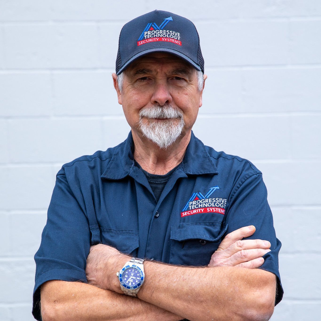 Man in navy hat and shirt with crossed arms, standing against a white wall.