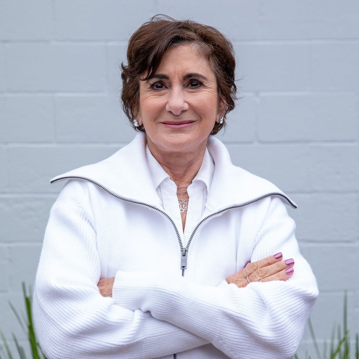 Woman with crossed arms, wearing a white jacket, in front of a light gray brick wall.