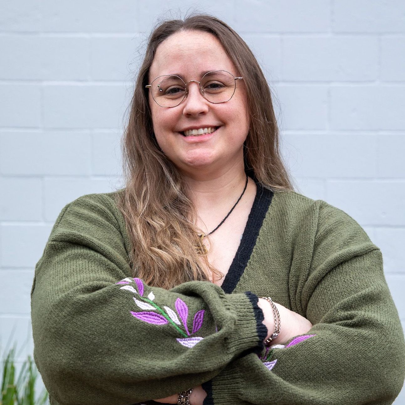 Woman with glasses smiles, arms crossed, wearing olive sweater with purple embroidery, standing outside.