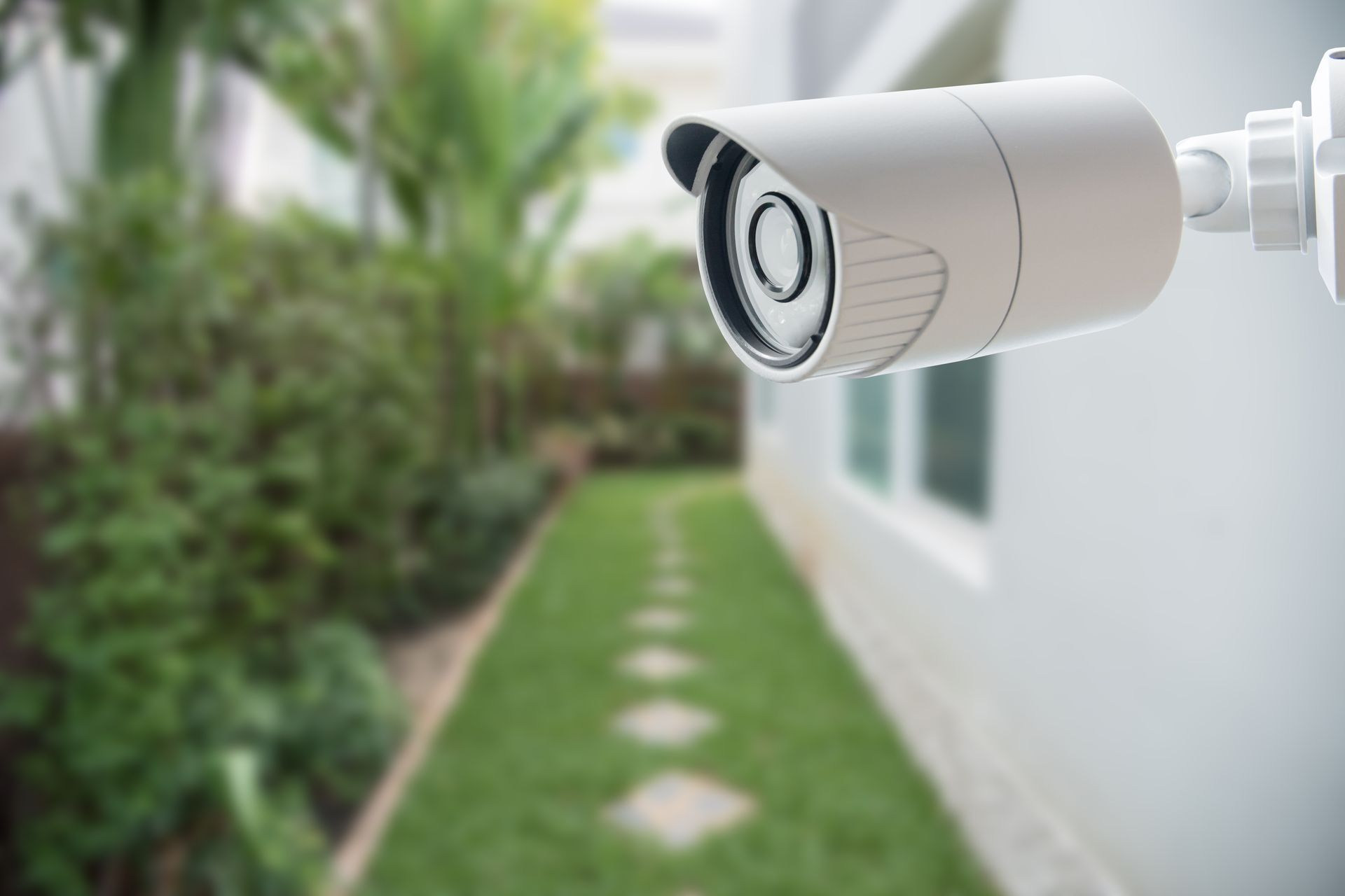 Security camera mounted on white building, overlooking backyard pathway.