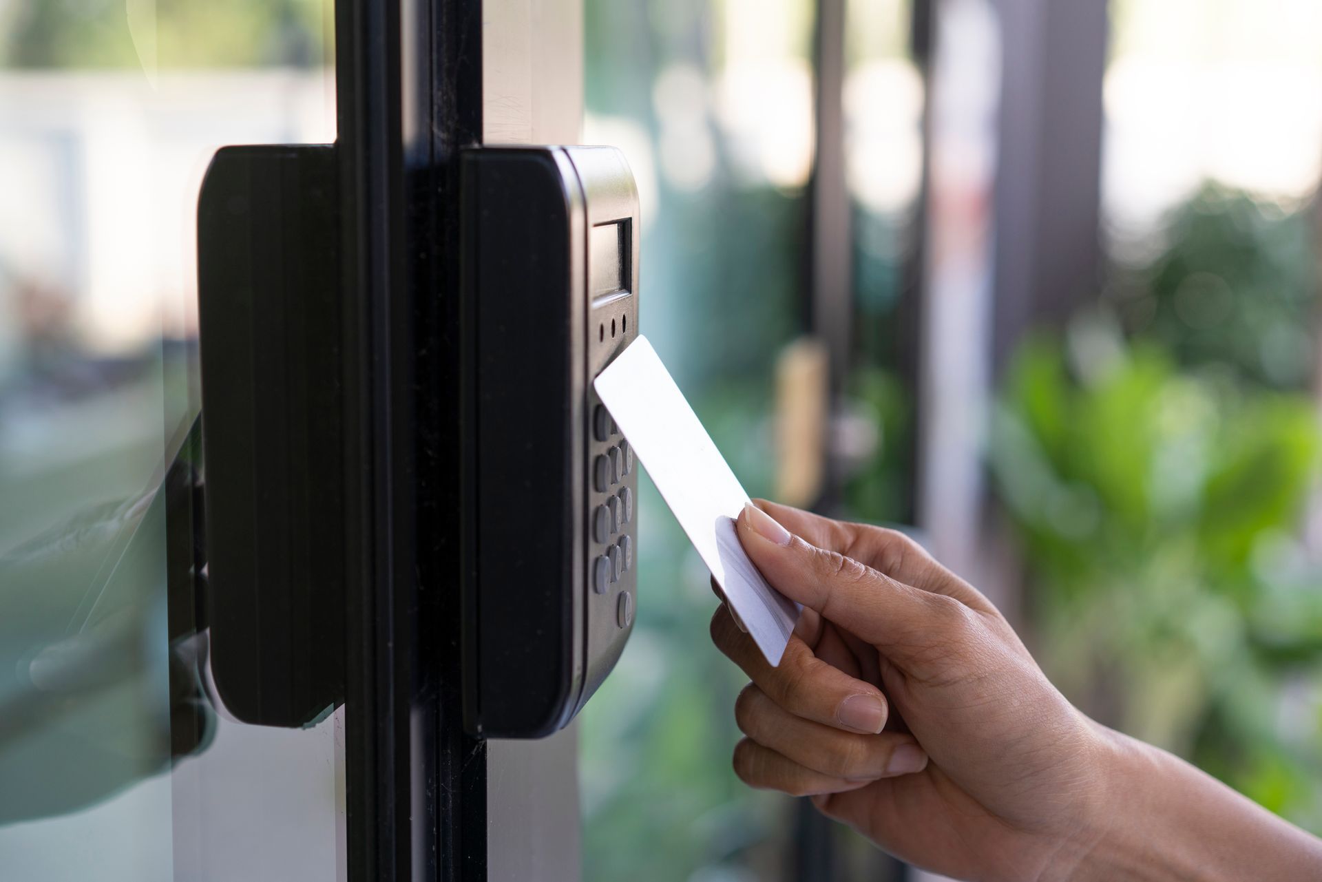 Person swipes a card at a black door access system on a glass door; green foliage visible in the background.