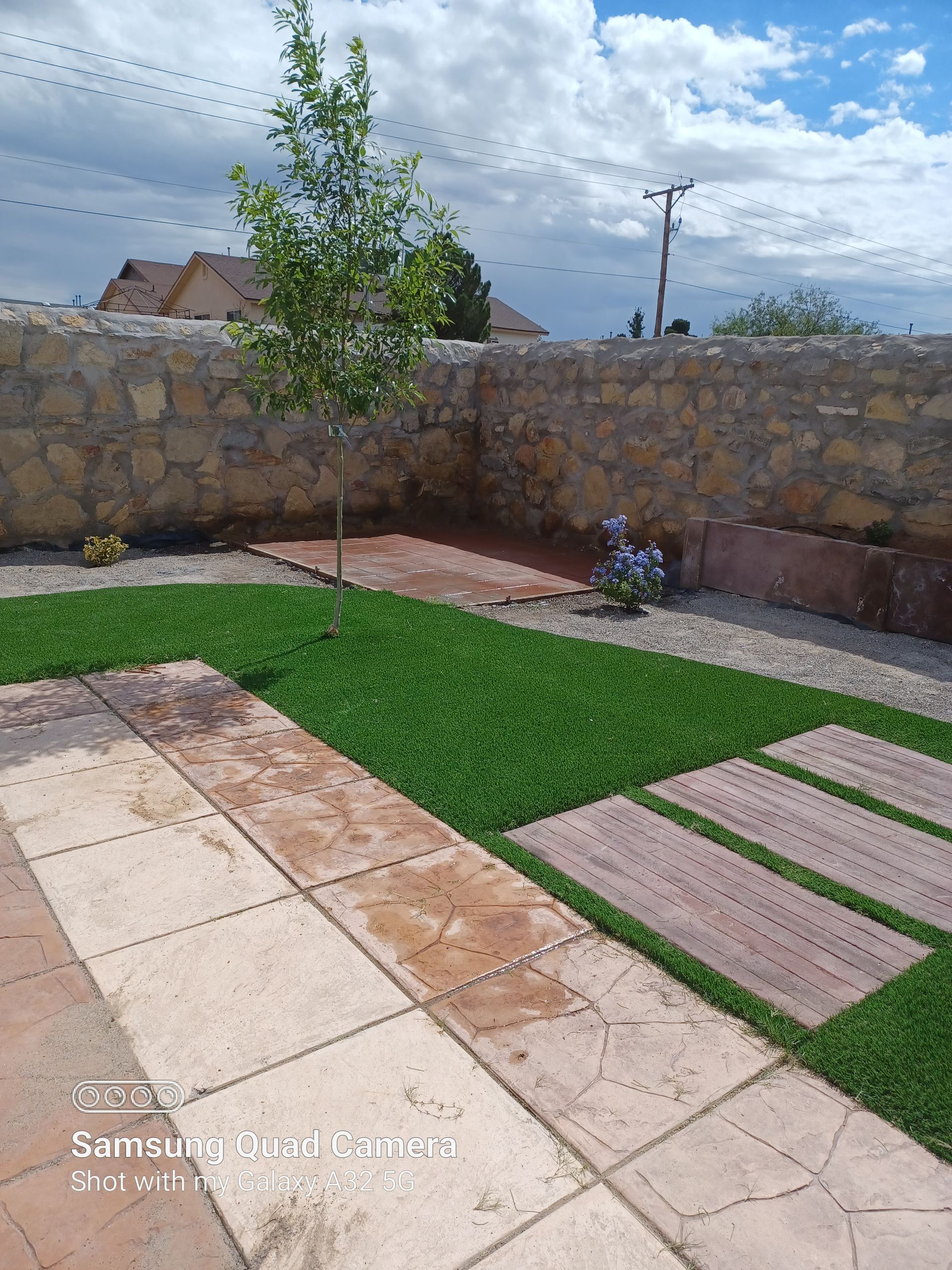 Stone patio and path with green grass and a small tree against a stone wall.