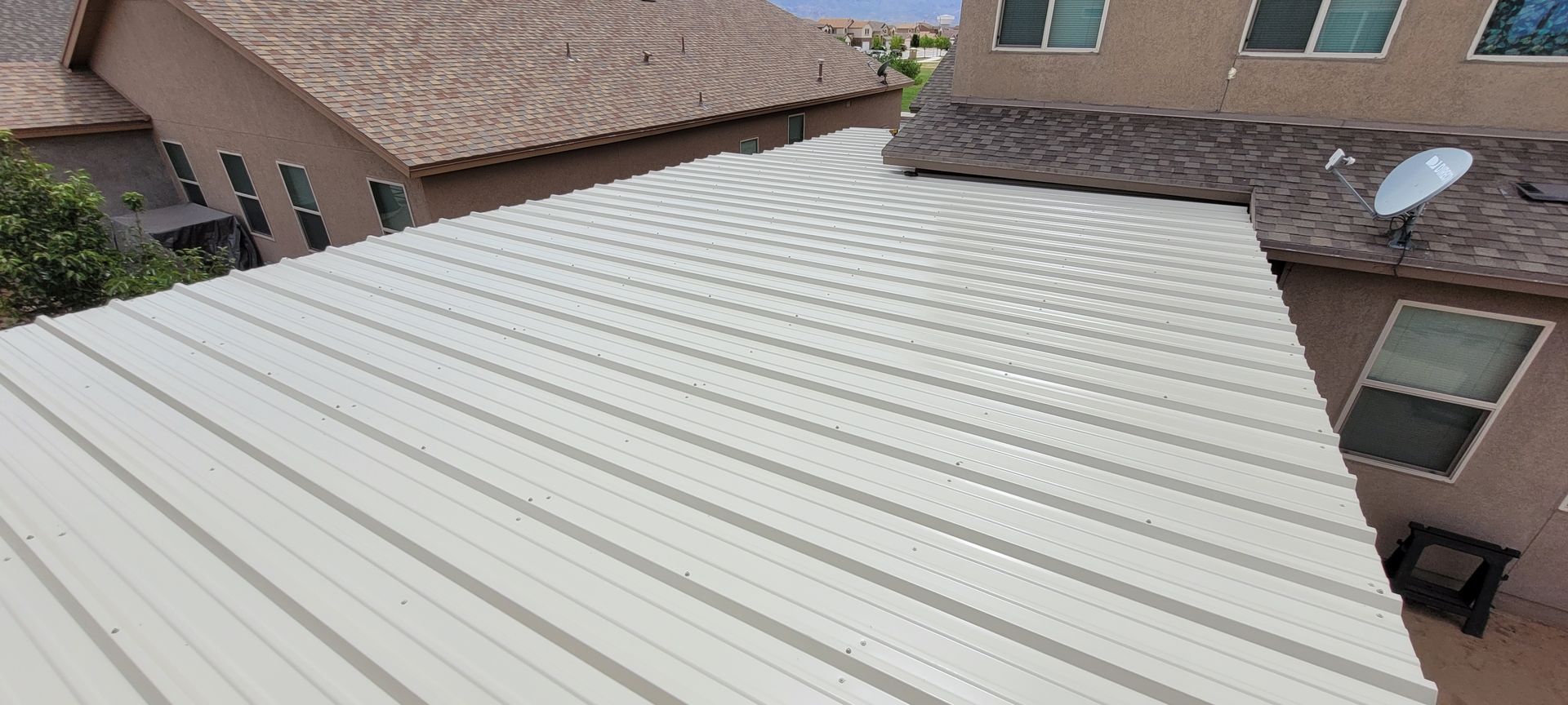 A view of a white metal roof next to a brown shingled roof, in an outdoor residential setting.
