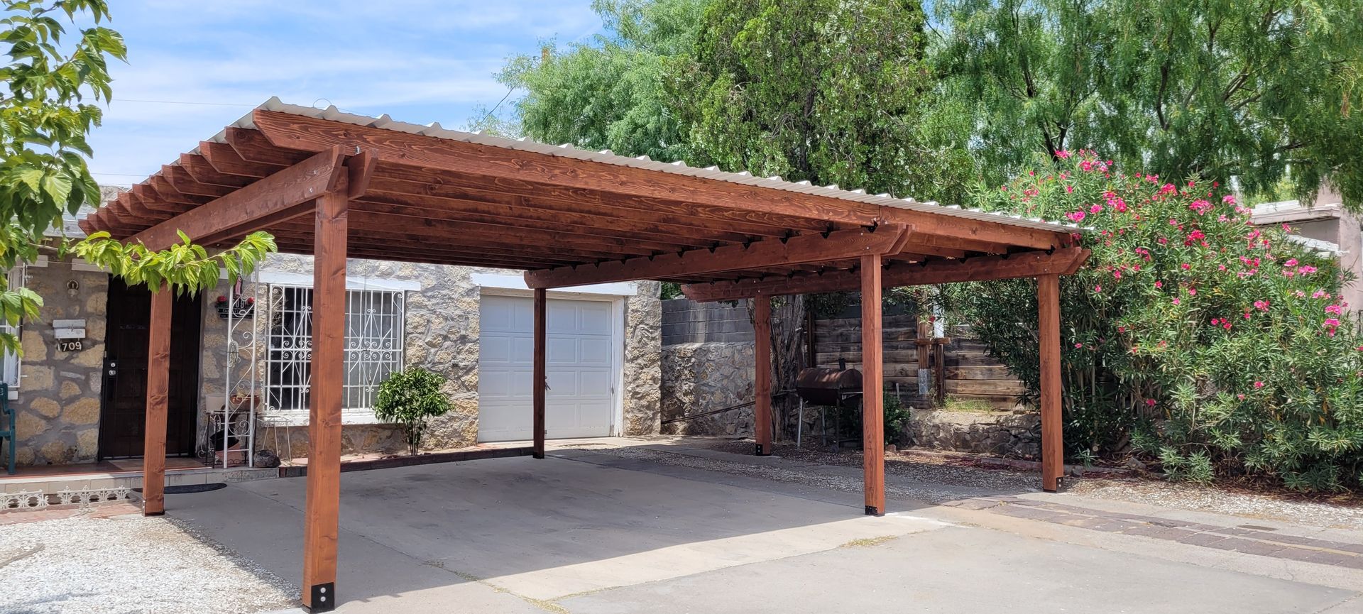 A brown wooden carport covers a driveway in front of a stone house with green trees in the background.