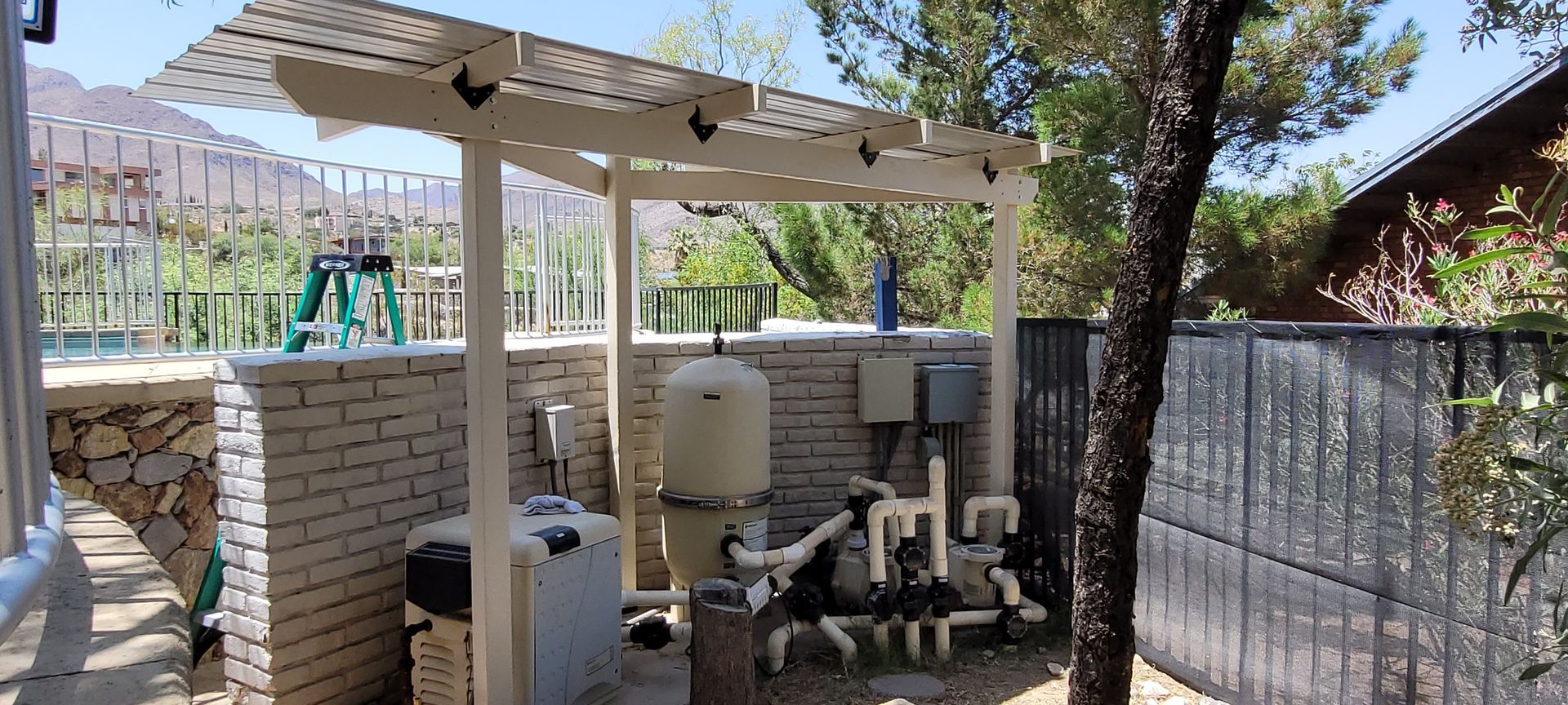 Pool equipment covered by a pergola. Brick wall on the left and a fence and tree on the right. Mountains in the background.