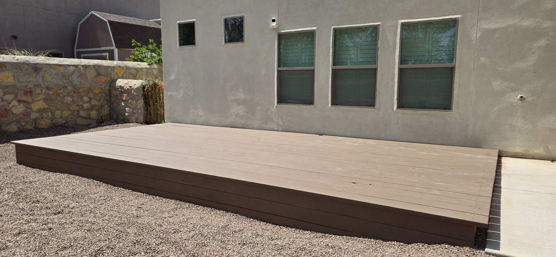 A composite deck in a backyard next to a building with windows and a gravel area.