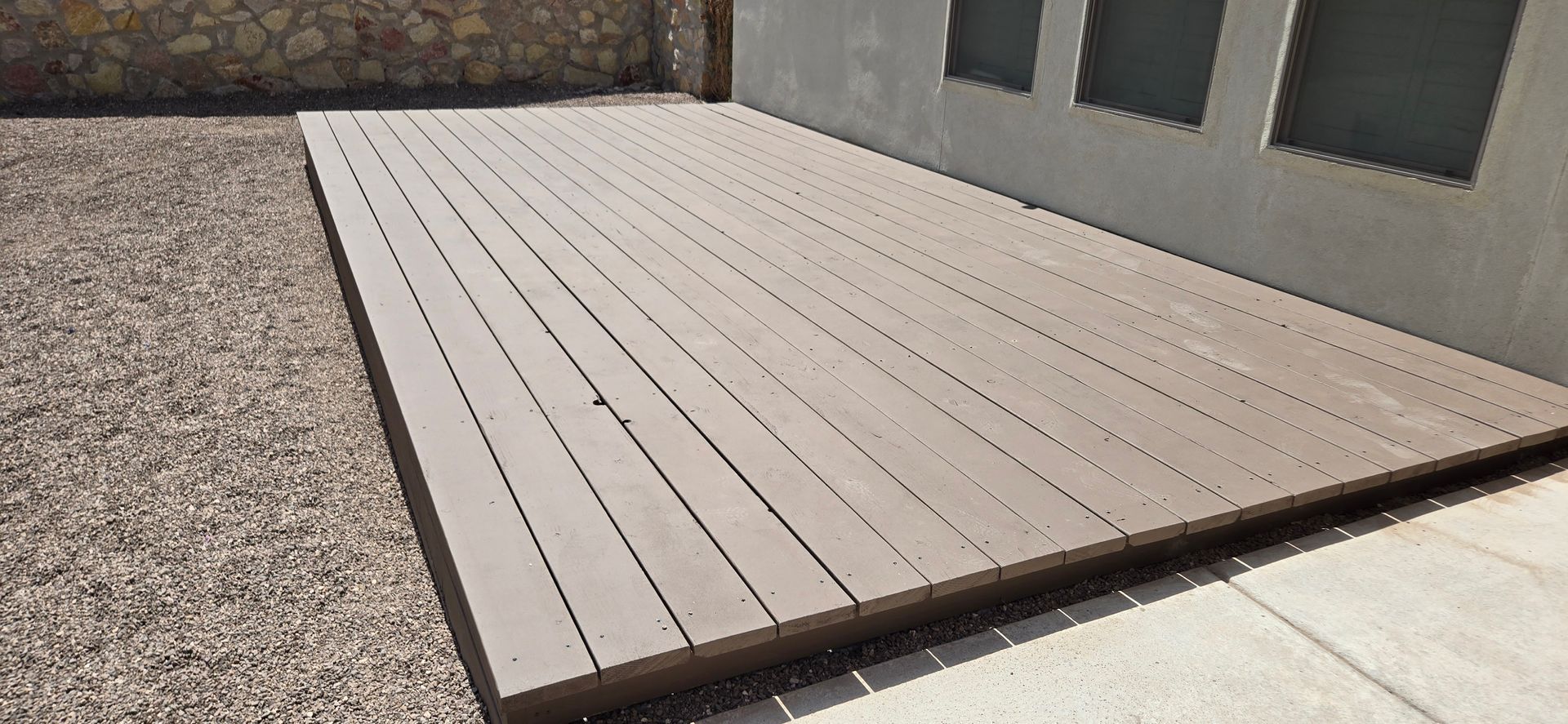 Wooden deck next to gravel and a wall with three small windows.