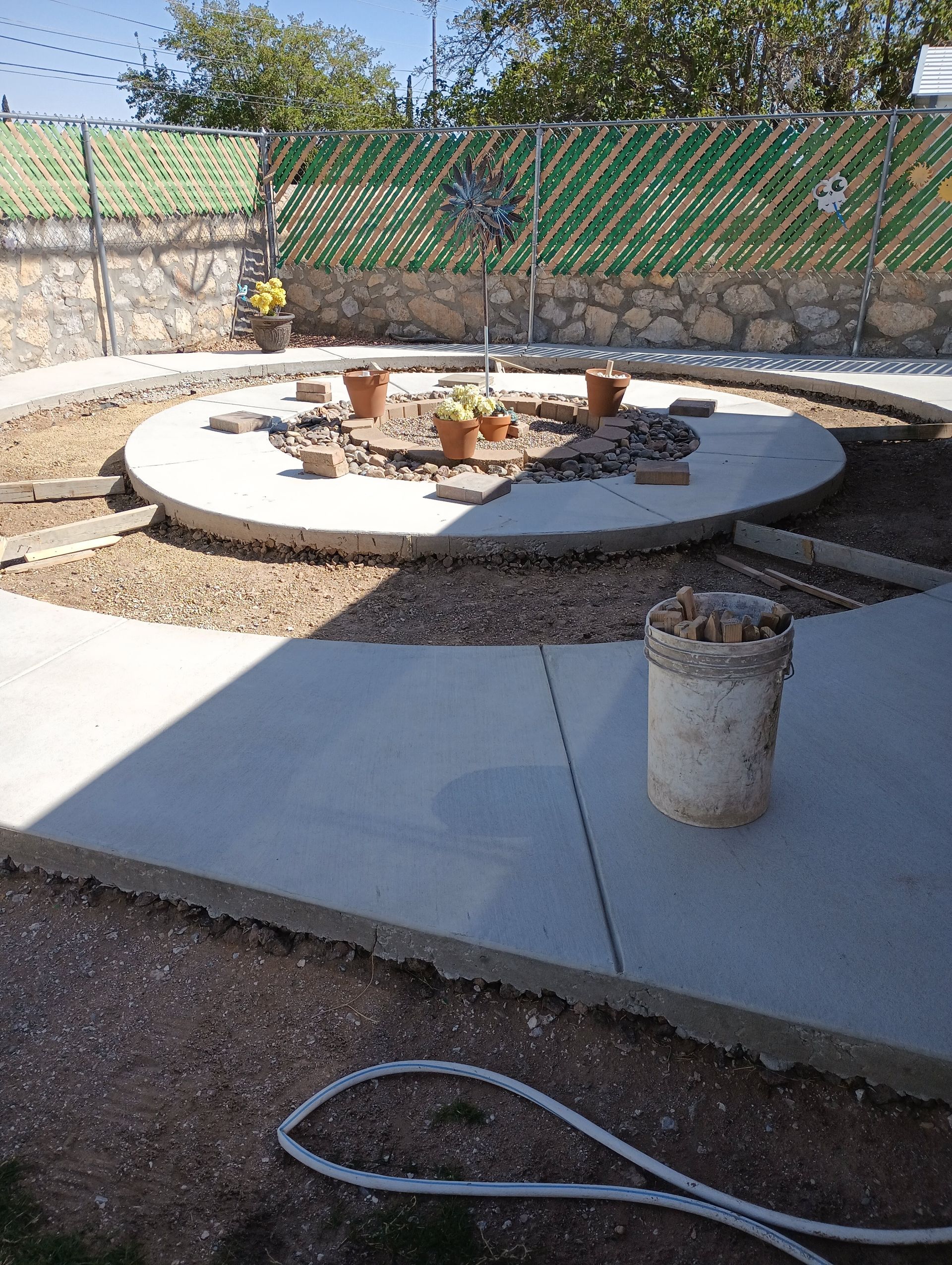 Concrete patio under construction with a circular center, several potted plants, and a brick wall in the background.