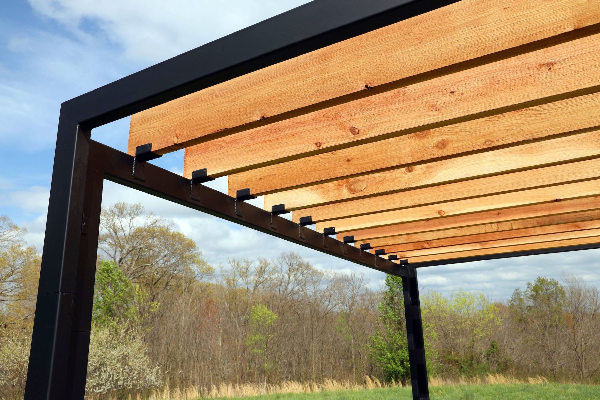 Black metal pergola with wooden slats, outdoors with a blue sky and trees.