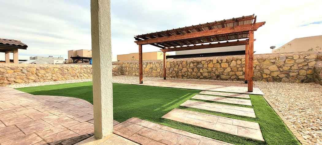 A backyard with a stone patio, a pergola, and green grass. A stone wall borders the yard.