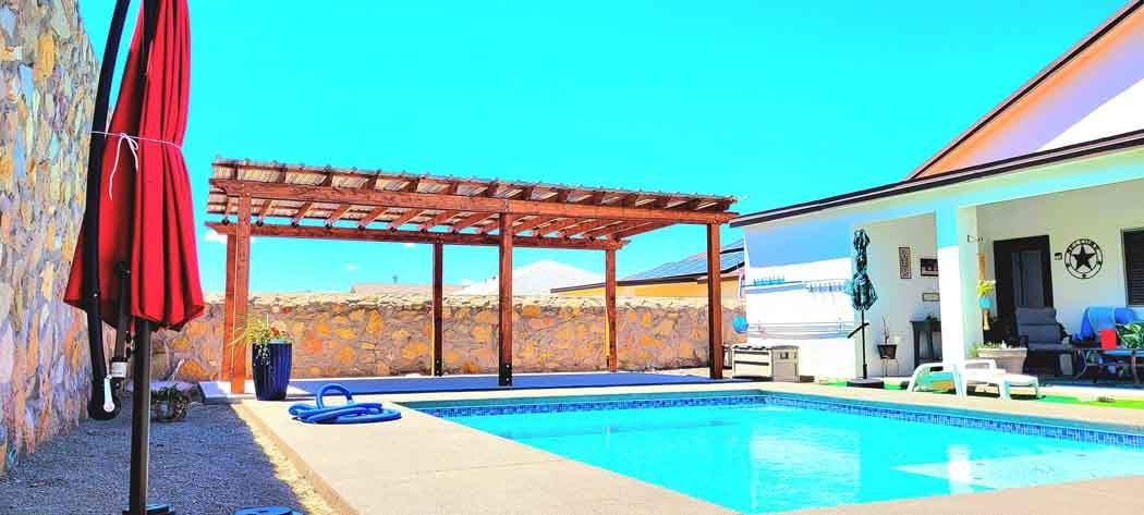 Poolside view with a red umbrella, wooden pergola, and clear blue sky.