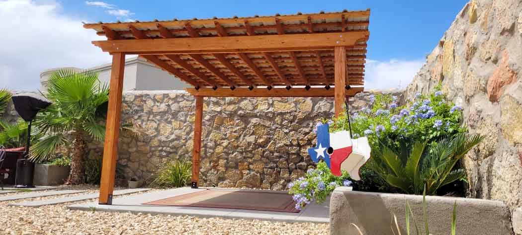 Wooden pergola in a yard, Texas flag, blue sky, stone wall, and plants.