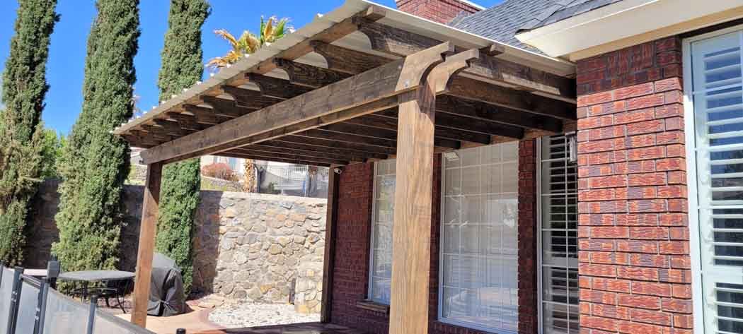 Wooden pergola over a brick building entrance. Tall green trees and blue sky in the background.
