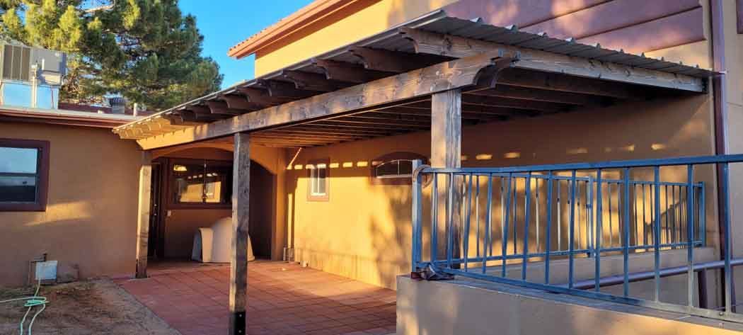 A covered patio with wooden beams, posts, and a brick floor. Sunlight casts long shadows.