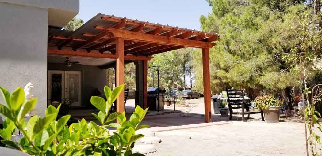 Wooden pergola extending from a building, over a patio. Lush foliage surrounds the area.