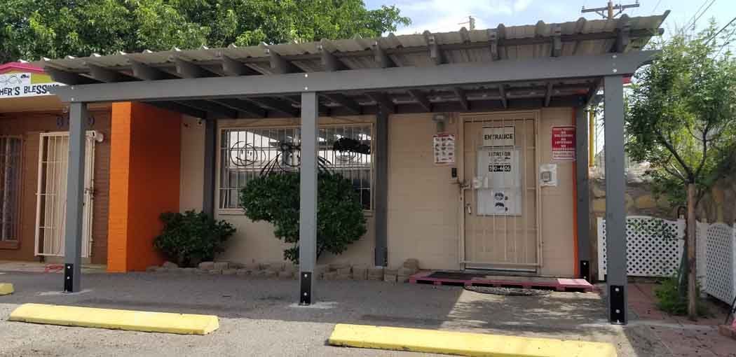 A low, gray pergola over a tan building with an orange wall and a closed door. Yellow parking curbs.