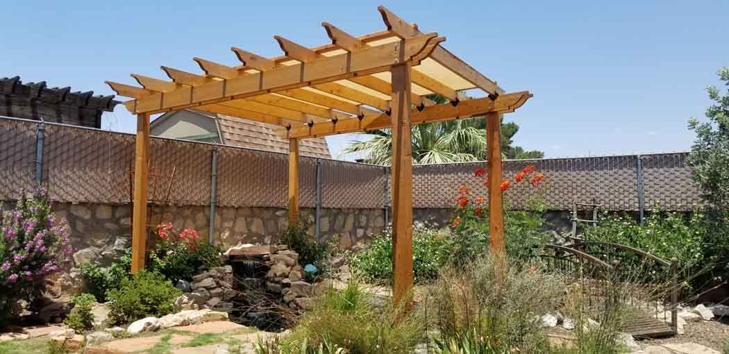 Wooden pergola in a garden with a rock wall and various plants under a blue sky.