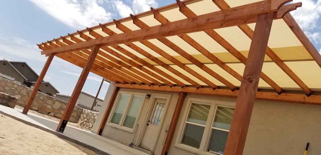 Wooden pergola with beige canopy over a house patio. Blue sky in the background.
