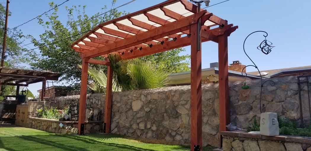 Wooden pergola archway in a sunny outdoor setting with a stone wall and a grassy lawn.