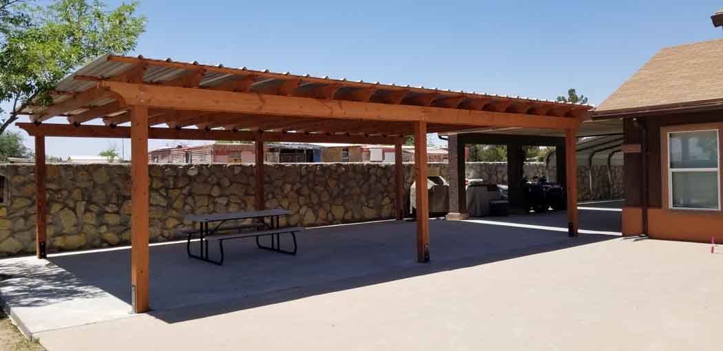 A brown wooden carport in a backyard, with a picnic table and a car parked inside another carport.