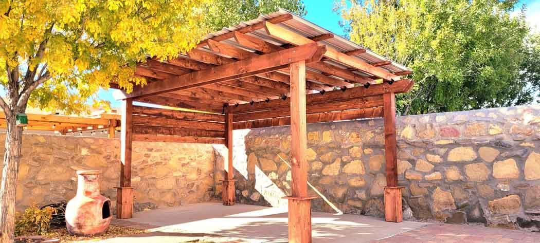 Wooden pergola in a yard with stone walls and foliage.
