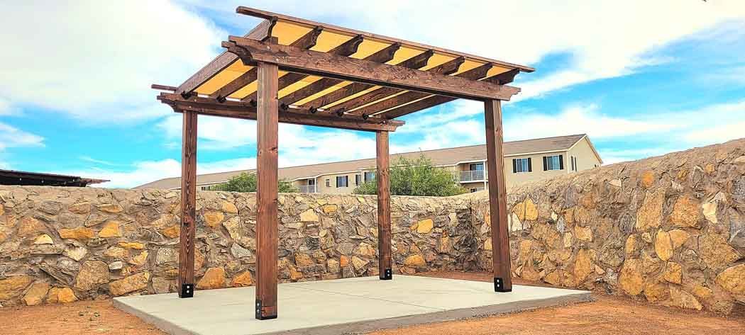 Wooden pergola on a concrete pad, set against a stone wall, with a blue sky background.