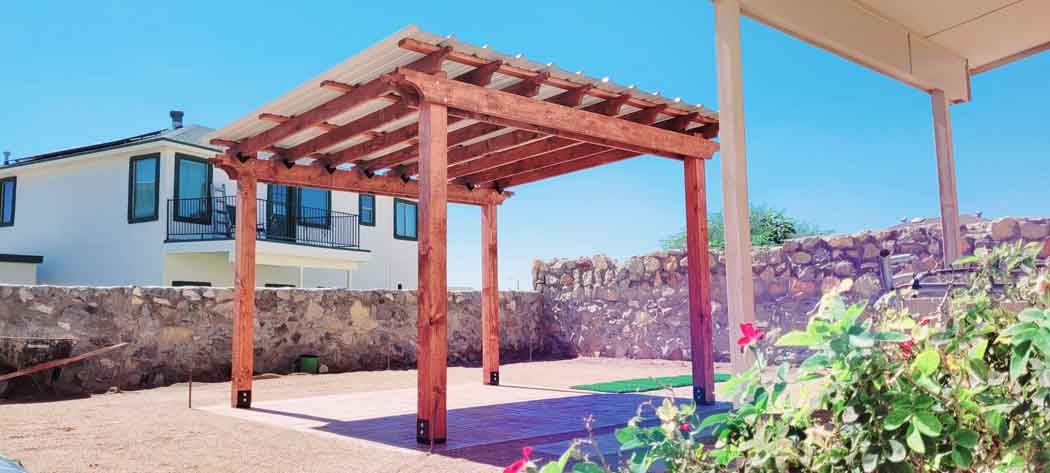 A wooden pergola in a backyard with a blue sky. A house is in the background, and greenery in the foreground.