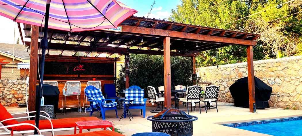 A backyard patio with a swimming pool, lounge chairs, and a wooden pergola. Various chairs are present.