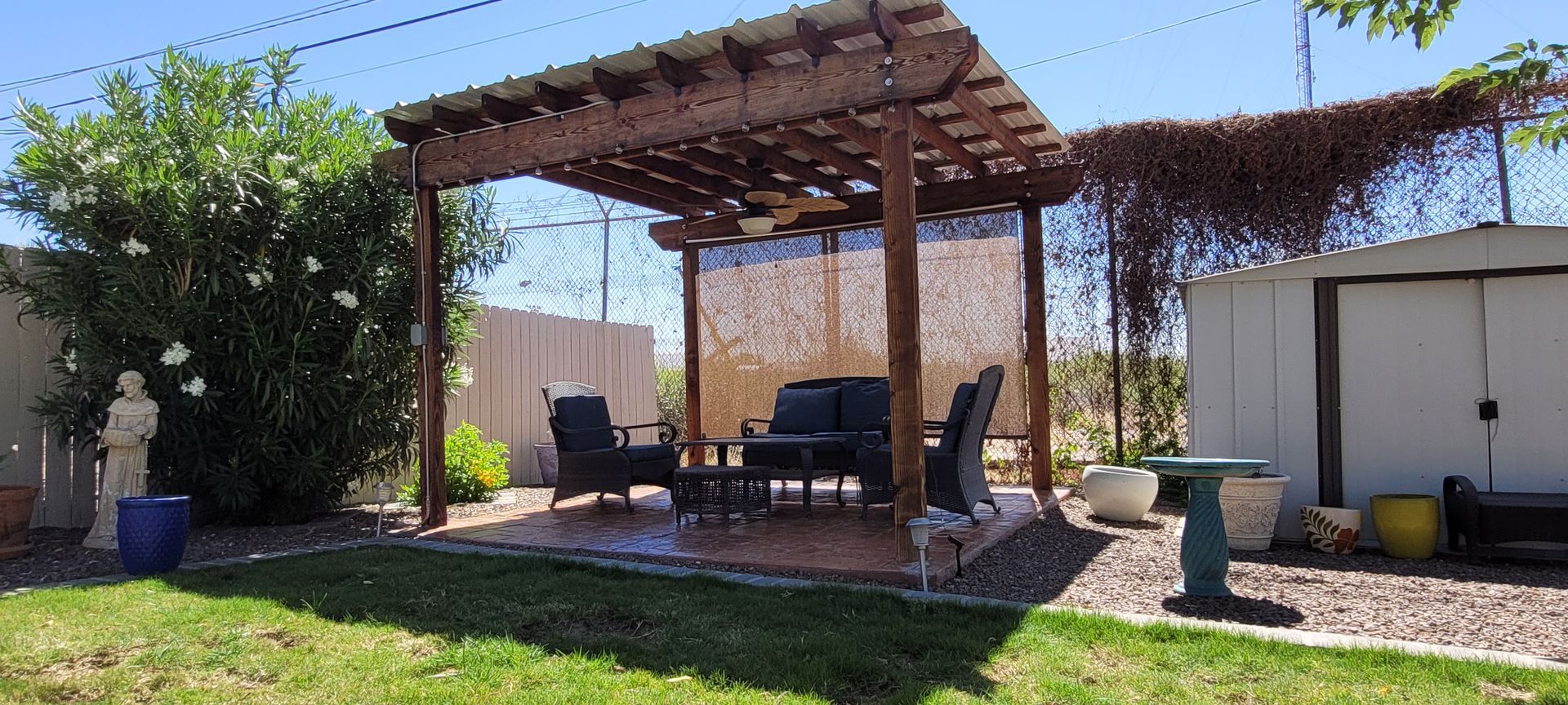 A backyard patio with a wooden pergola, seating, and a small storage shed. Green grass, blue sky.