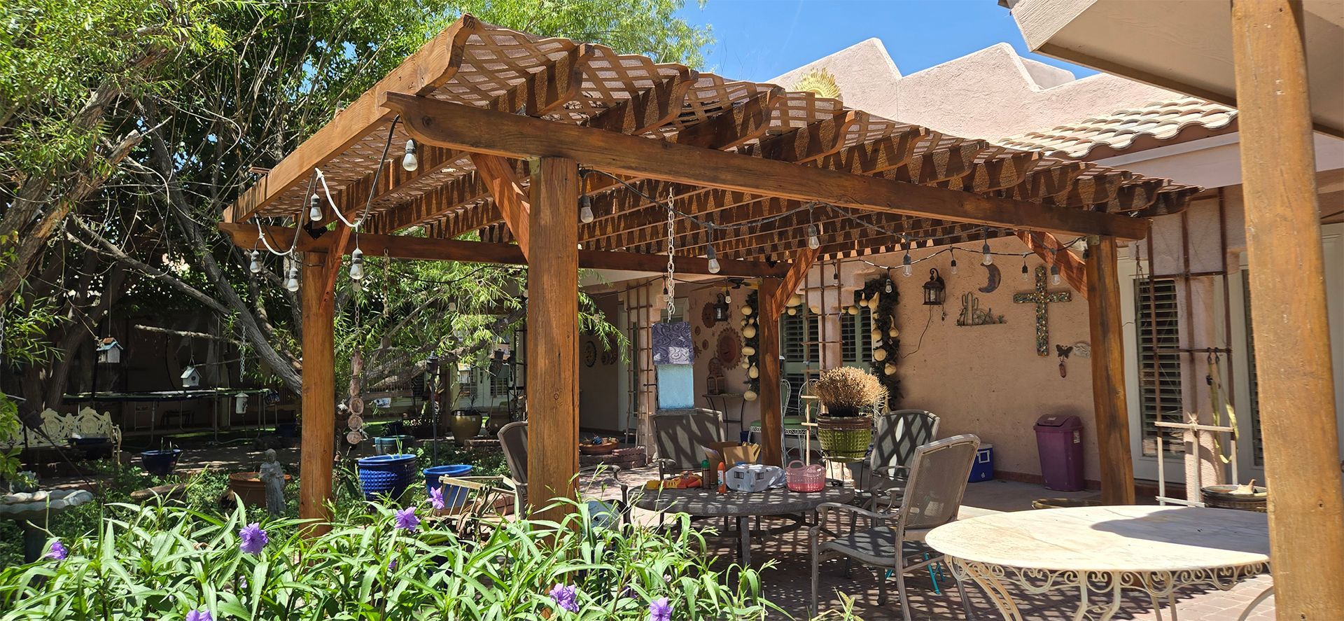 Wooden pergola over a patio, with table and chairs. Green plants, blue sky, and a house in the background.