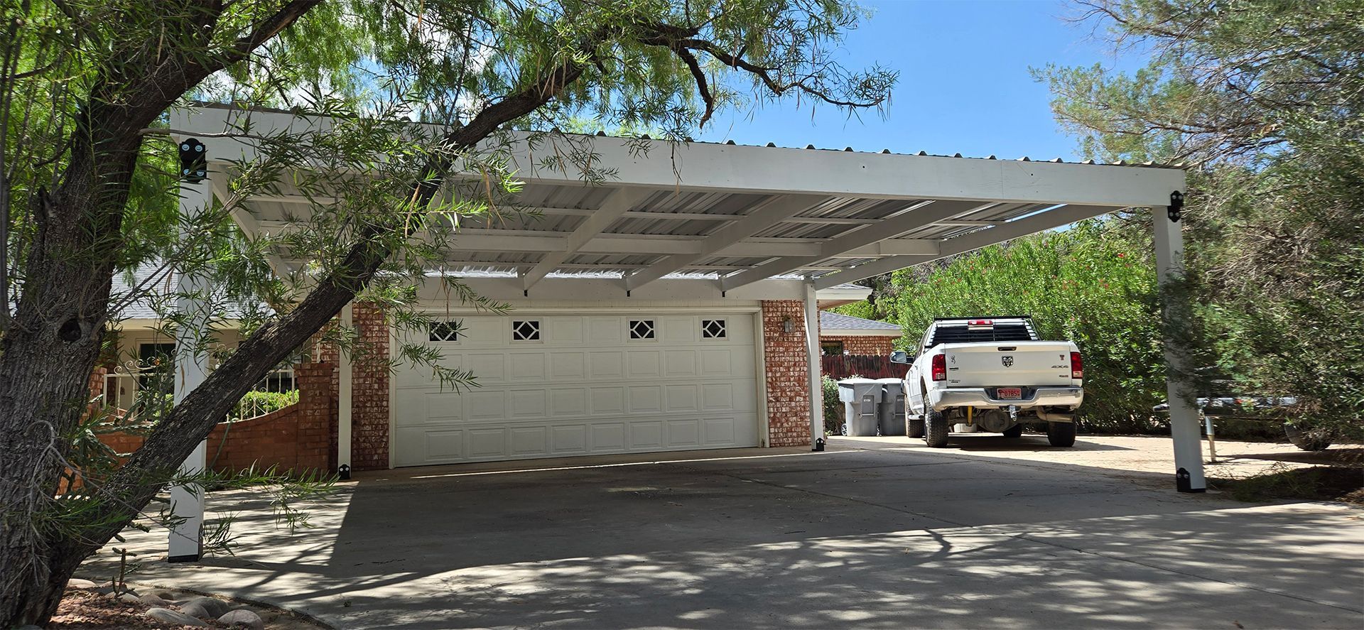 White pickup truck parked under a carport in front of a white garage, a tree is in the foreground.