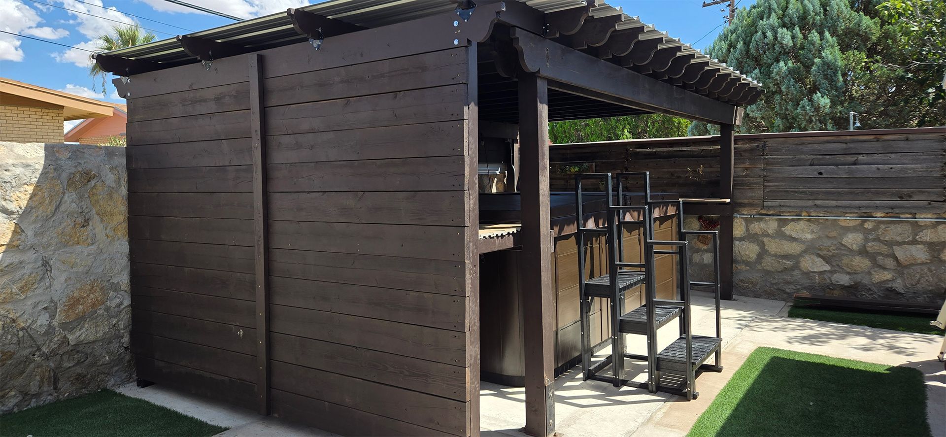 Wooden outdoor structure with a dark brown exterior, partial roof, and steps, set against a stone wall.