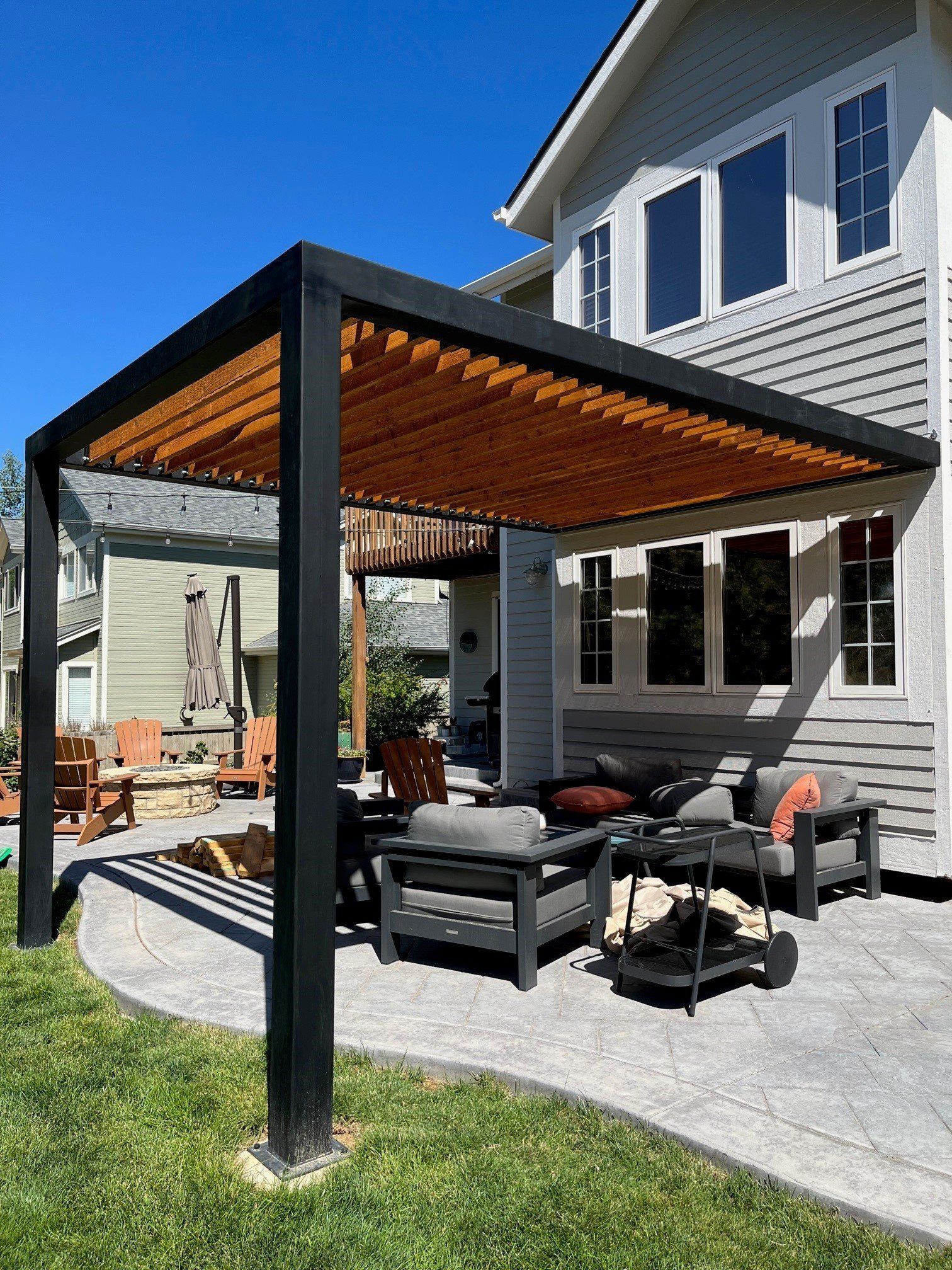 Black pergola with wooden slats over patio seating area; sunny backyard.