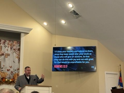 A person in a suit speaks from a podium in a church, with a screen behind them displaying Hebrews 13:17.