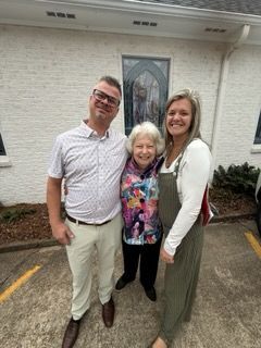 A man, a woman in a colorful jacket, and a woman in a white shirt and green overalls standing together outside a building.