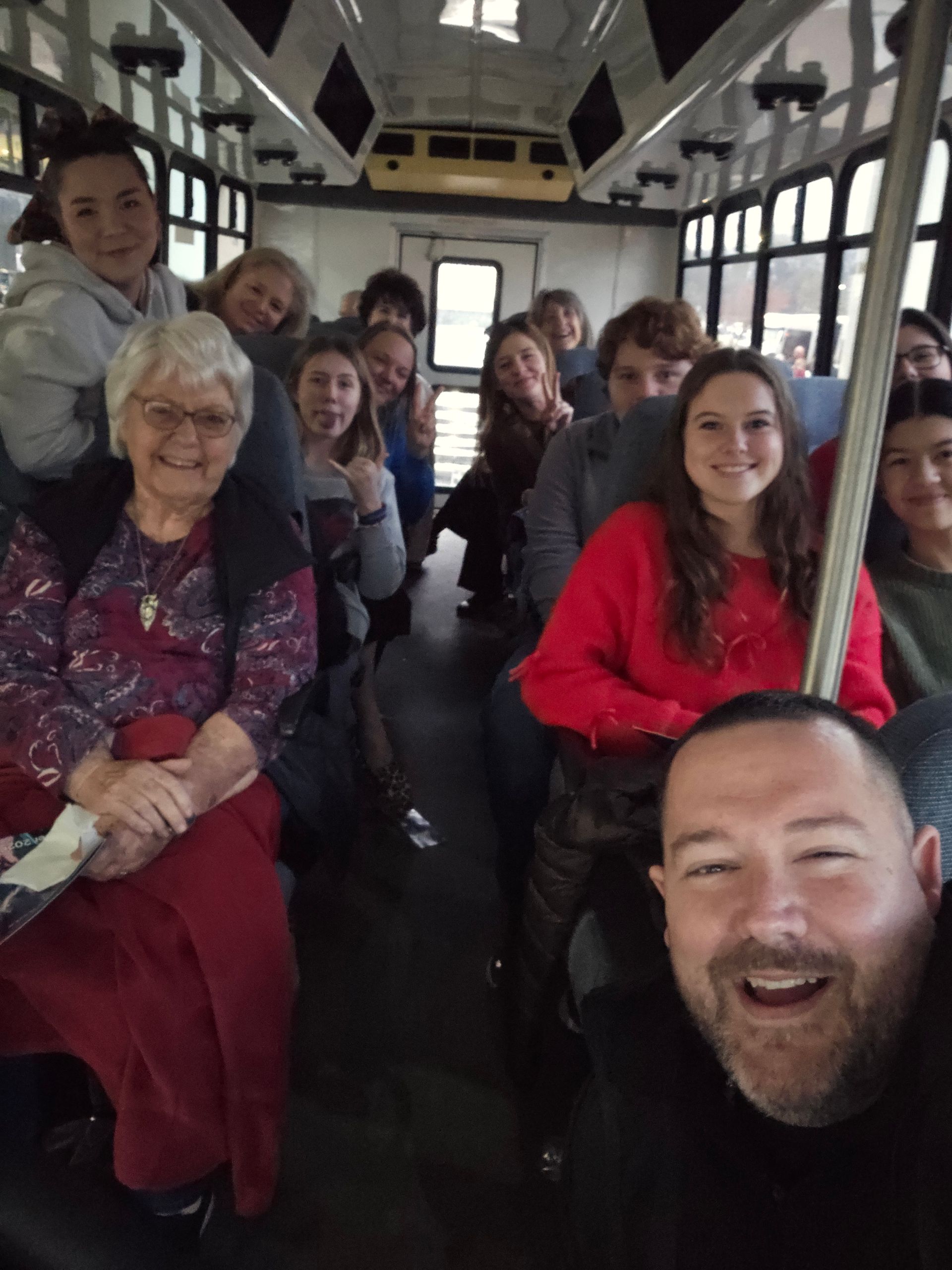 A group of people sitting inside a public transit vehicle, smiling for a selfie taken by a man in the foreground.