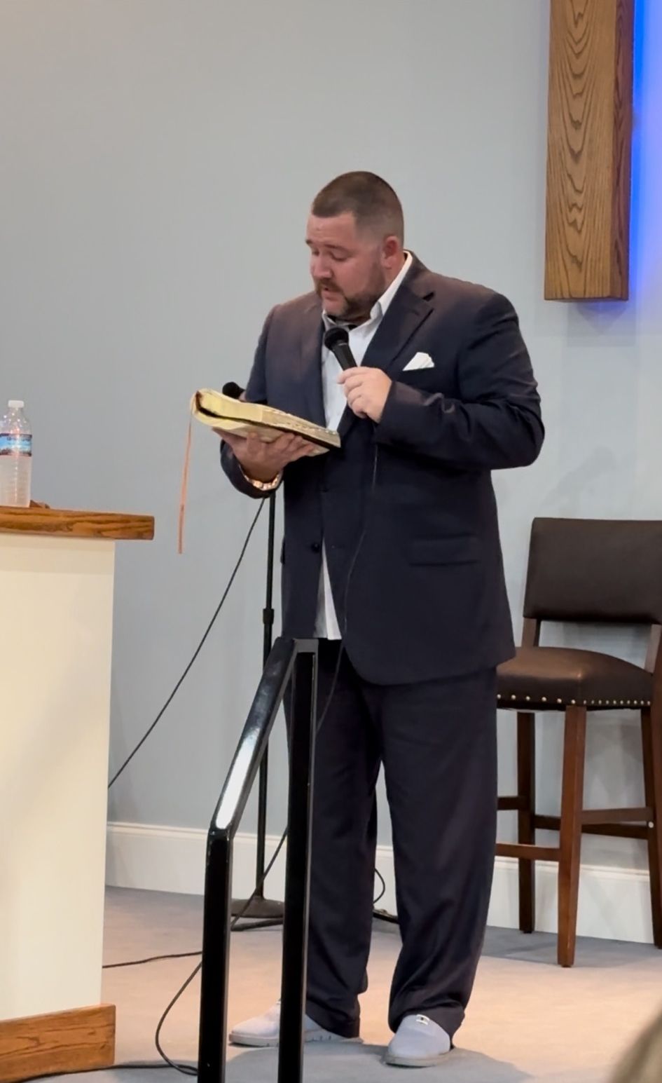 A person in a suit stands at a podium holding a book and a microphone, appearing to speak or read to an audience.