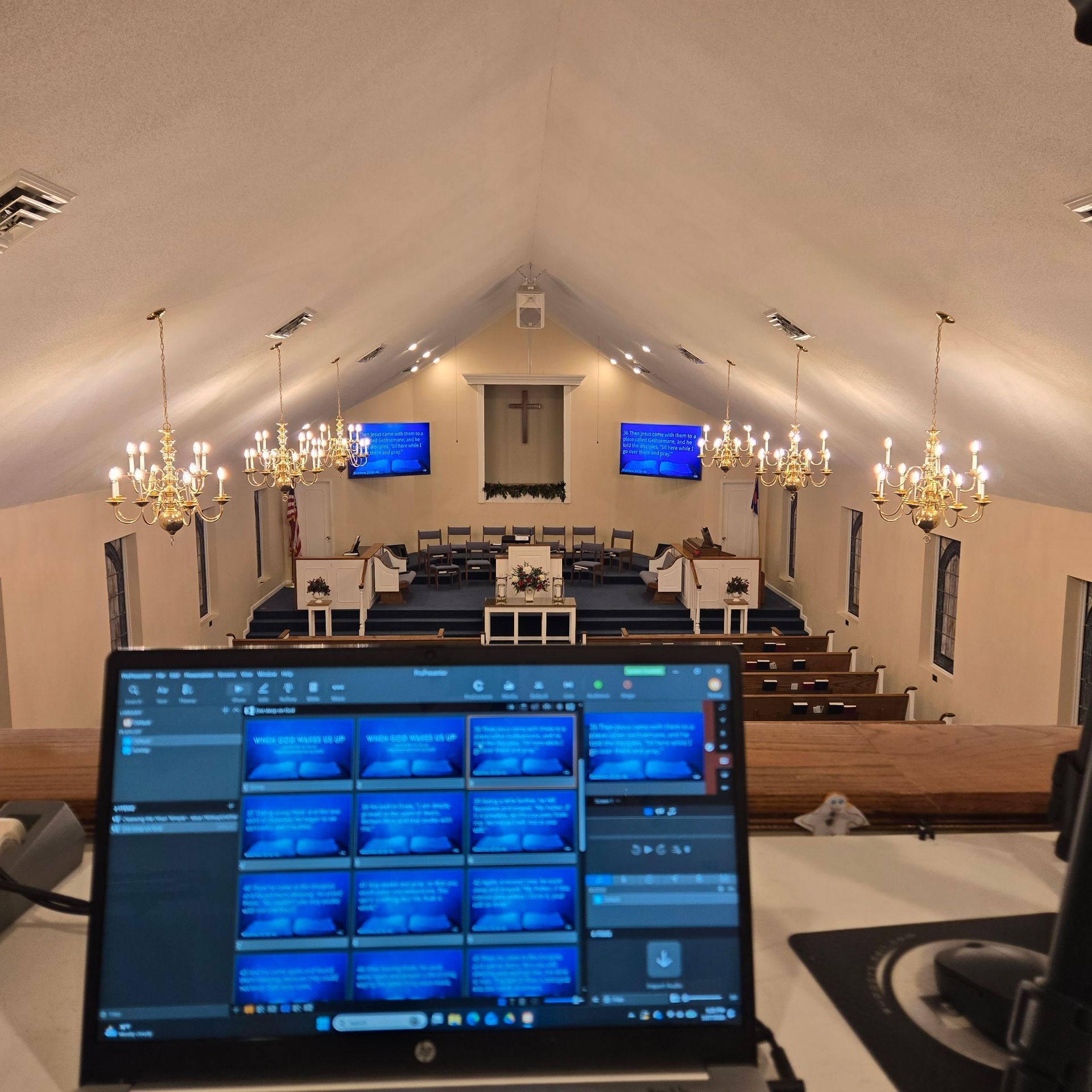 A laptop in a church loft shows video software, overlooking the sanctuary with a stage, two screens, and chandeliers.