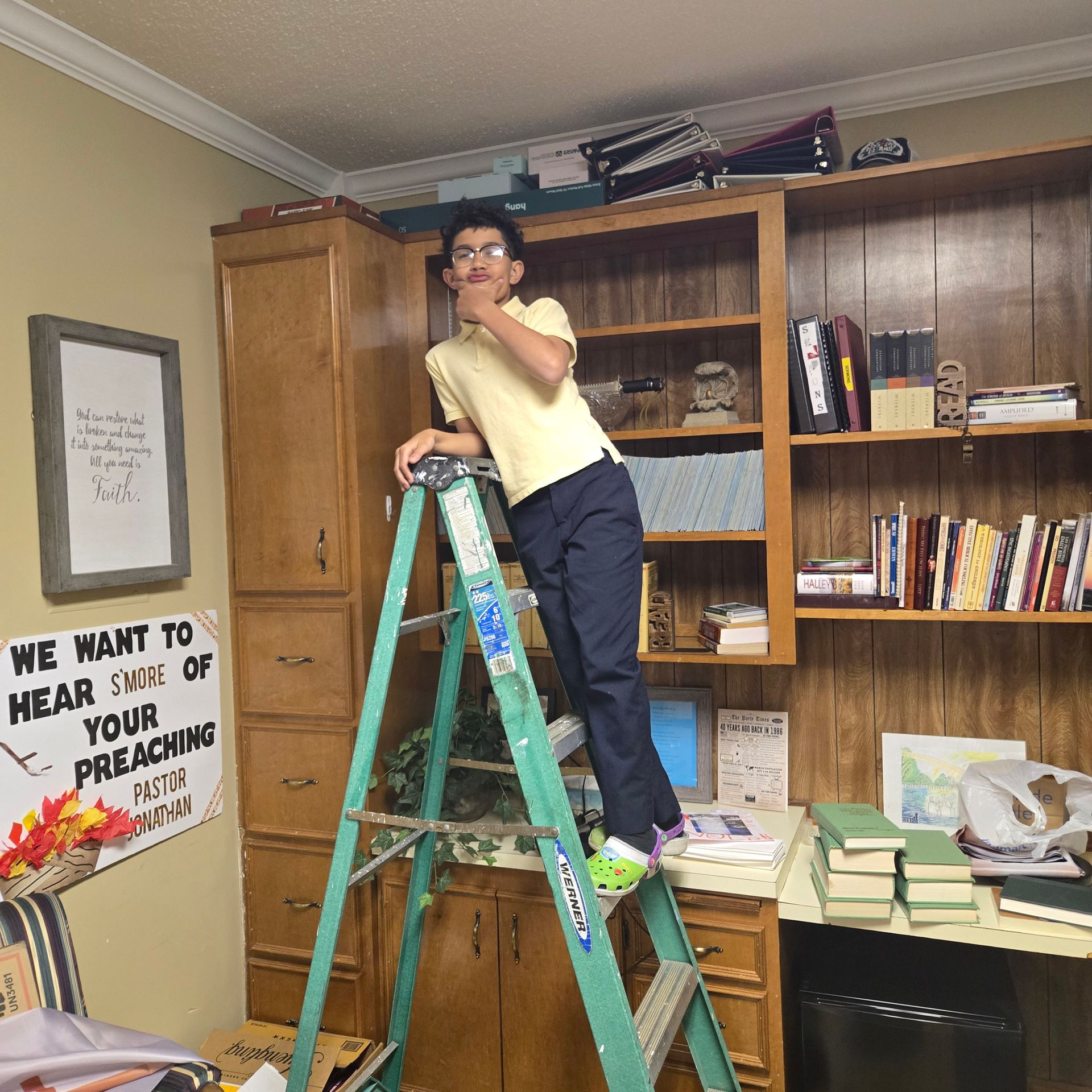 A person in a yellow polo shirt and dark pants stands on a green stepladder in front of a wooden bookshelf.