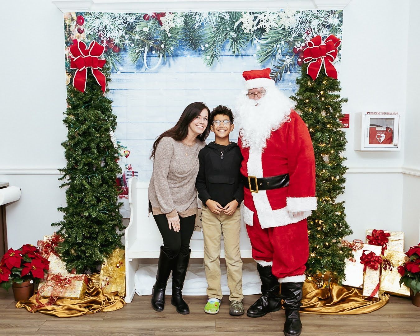 A smiling woman and child pose with Santa Claus in front of a festive backdrop with trees, bows, and wrapped gifts.