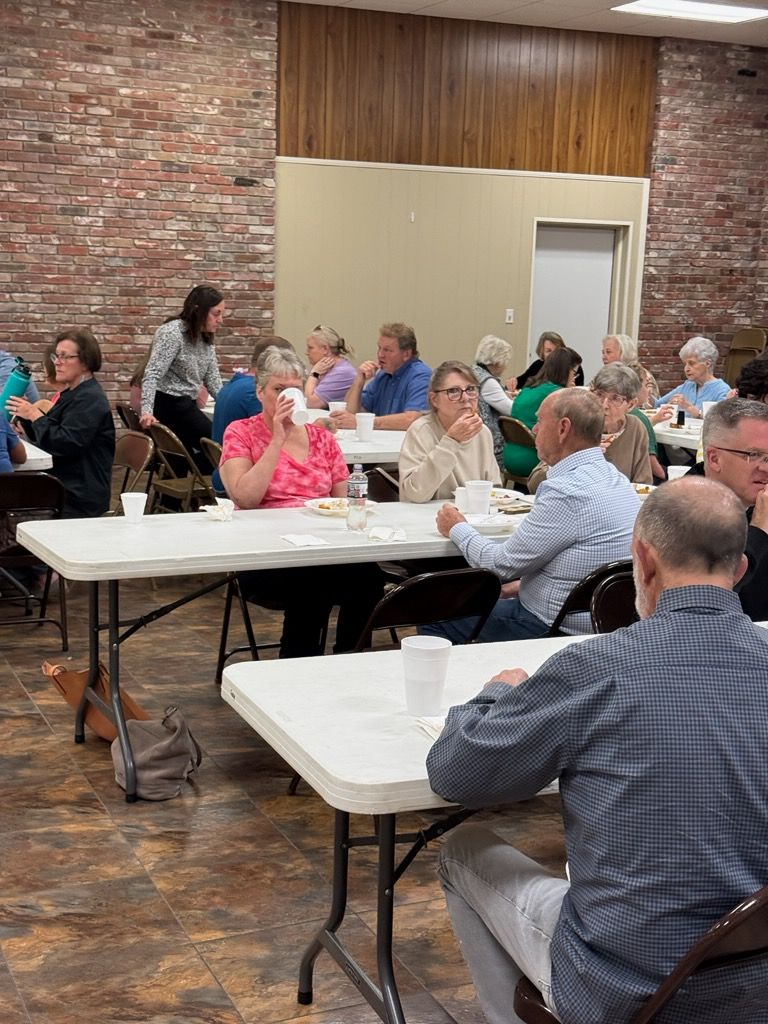 A group of people sit at white tables in a room with a brick wall, eating and talking during an indoor gathering.