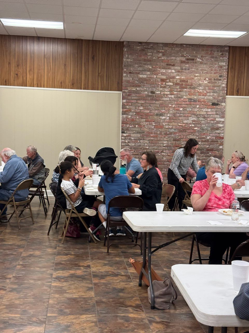People sit at tables in a room with a brick accent wall, eating and conversing during a gathering.