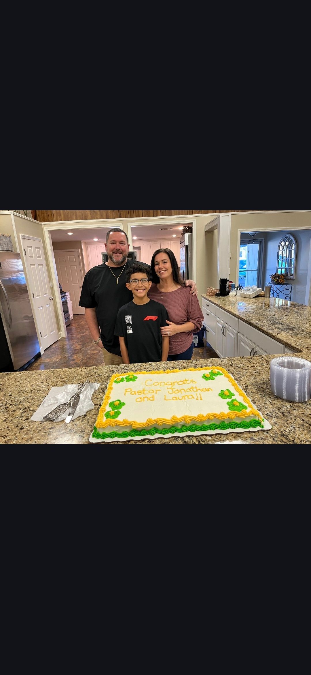 A smiling family of three stands arm-in-arm in a rustic banquet hall with stone walls and rows of white tables.