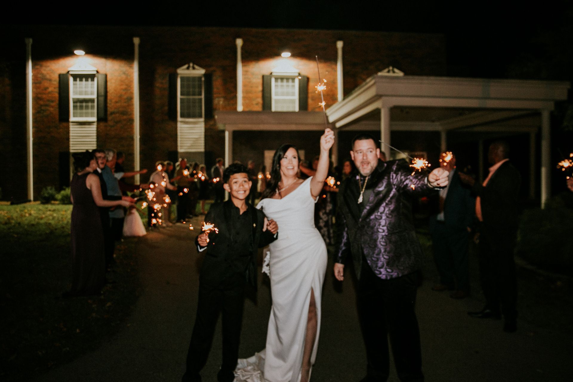 A bride and two people in formal attire hold sparklers outside a brick building at night during a wedding exit.