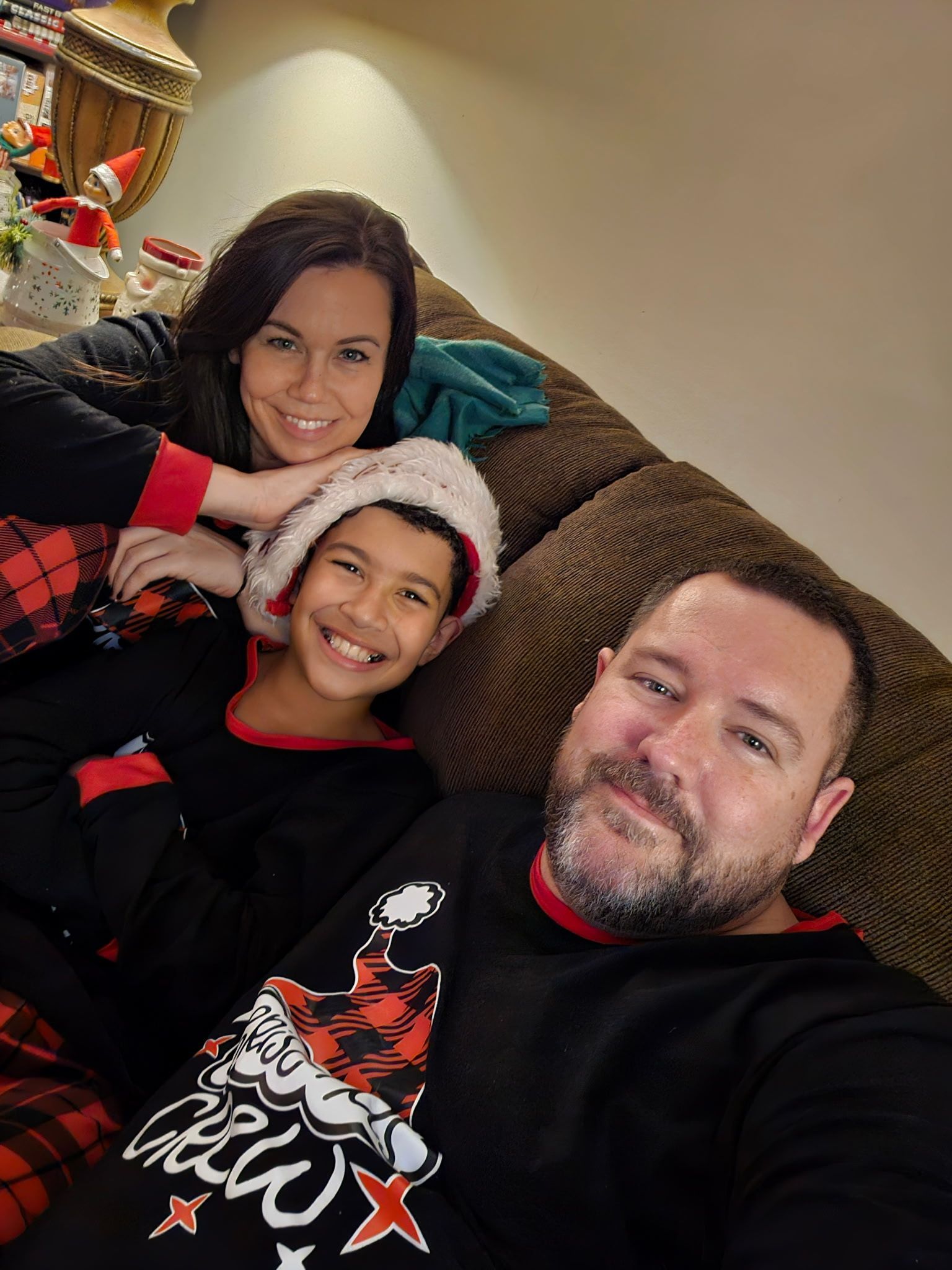 A family of three smiles for a selfie while wearing matching black holiday pajamas on a brown couch.