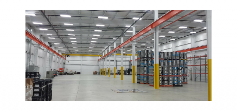 Interior of a large warehouse with stacked metal drums, overhead beams, and fluorescent lighting.