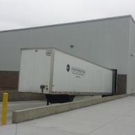 White semi-truck trailer backed into loading dock of a gray industrial building.